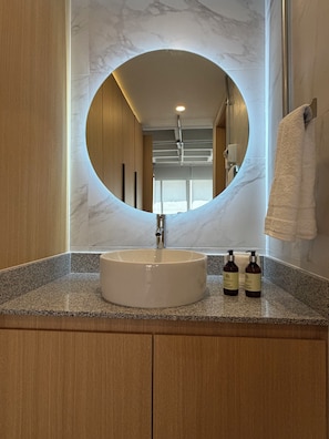 A bathroom with a round mirror, a sink, and two bottles of shampoo. - Modern 1BR in Juárez with fast WiFi, great natural light, a comfortable king bed, a fully equipped kitchen, and a luxury bathroom. Perfect for couples or solo travelers seeking comfort, style, and a prime location in Mexico City. (Cuauhtémoc)