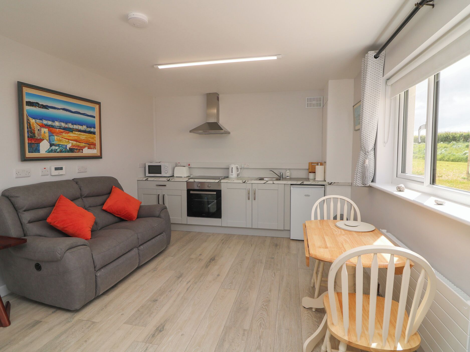 A compact living space with a kitchenette, a sofa with red cushions, a dining table with chairs, and a window with a view.
