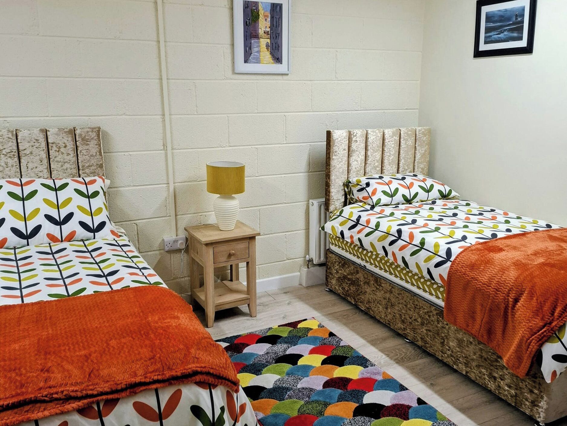 A room with two beds, a small wooden nightstand with a lamp, a colorful rug, and a framed picture on the wall.