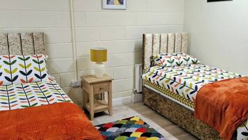 A room with two beds, a small wooden nightstand with a lamp, a colorful rug, and a framed picture on the wall.