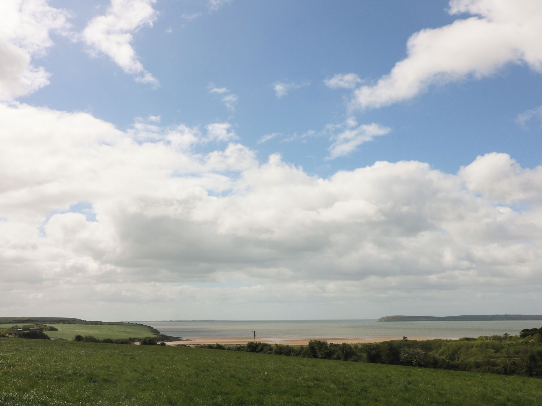 A coastal landscape with a beach, grassy fields, and a partly cloudy sky.