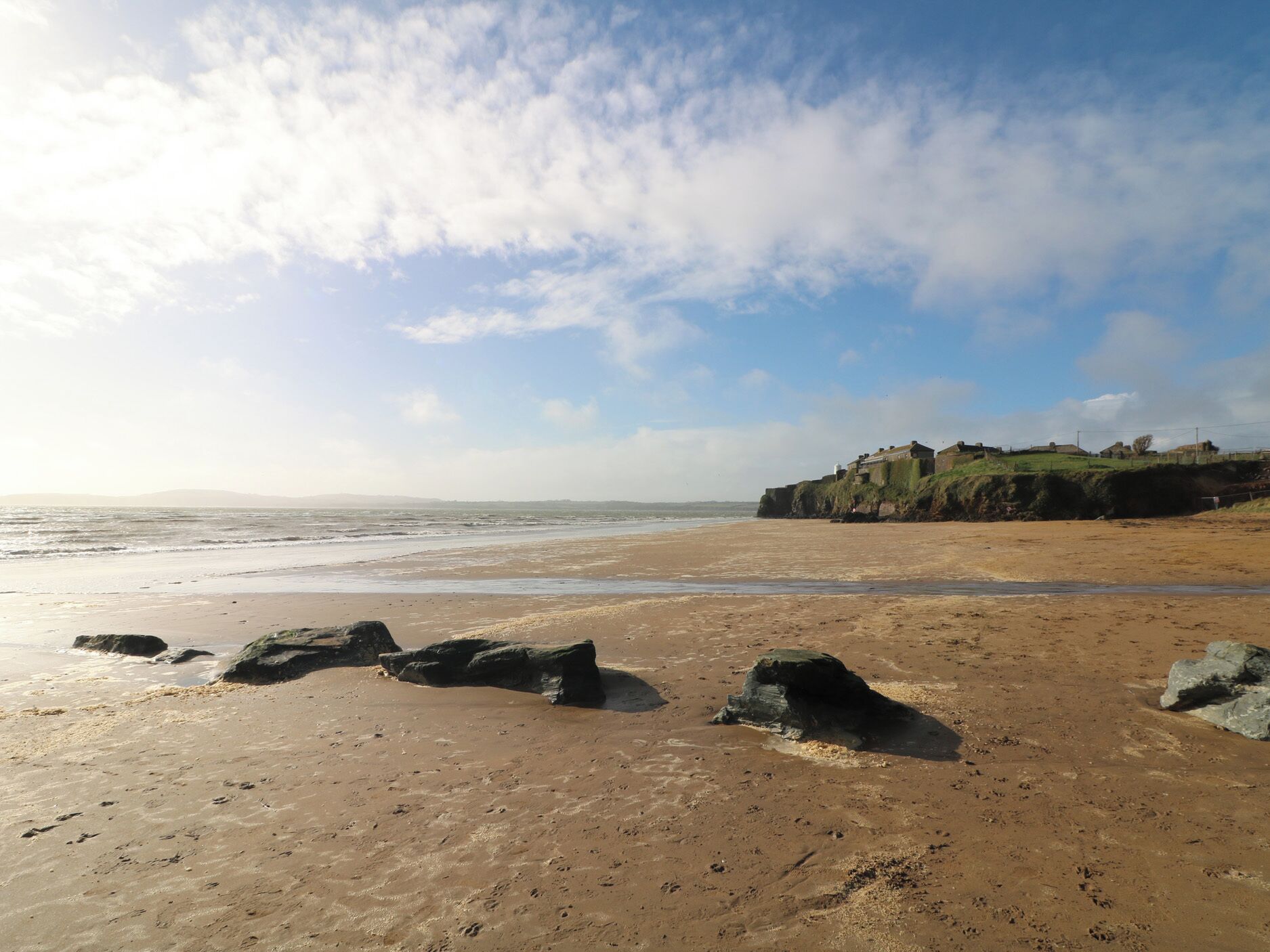 Une plage avec de gros rochers, une falaise sur laquelle se trouvent des bâtiments, et l’océan en arrière-plan.