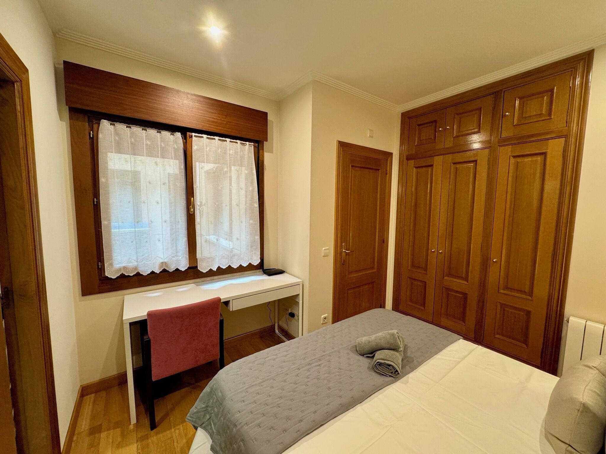 Un dormitorio con cama, escritorio con silla, ventana con cortinas y armarios de madera.