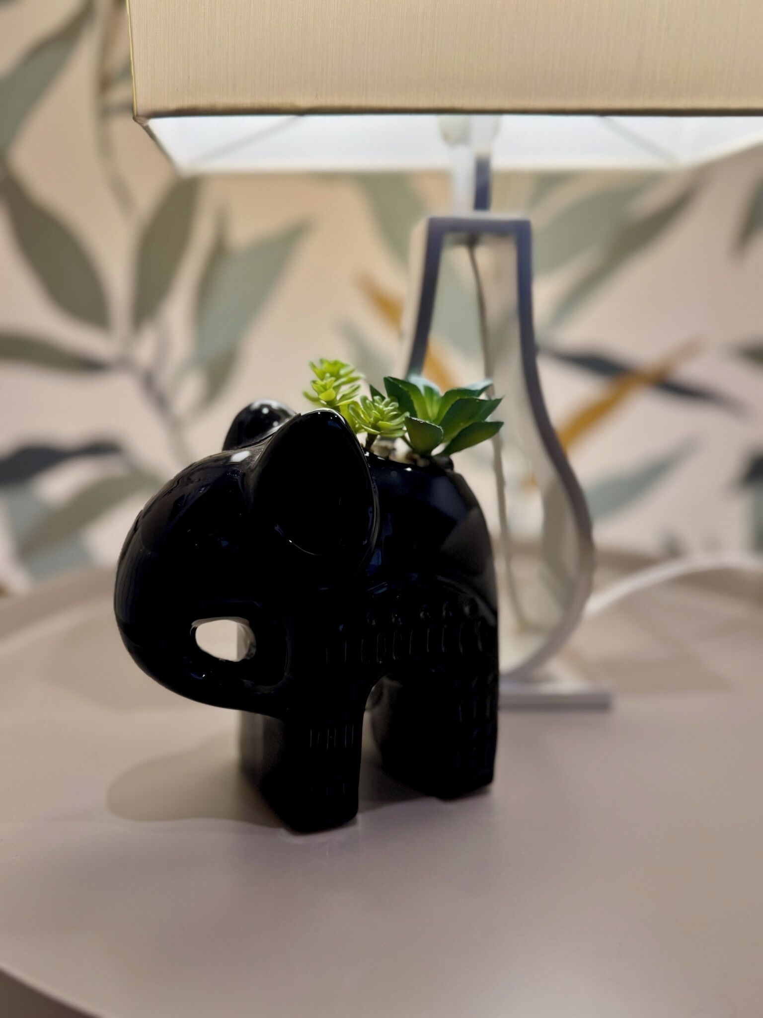 Eine schwarze, elefantenförmige Lampe mit einer kleinen Pflanze obenauf, platziert auf einer weißen Oberfläche, im Hintergrund ist ein Lampenschirm zu sehen.