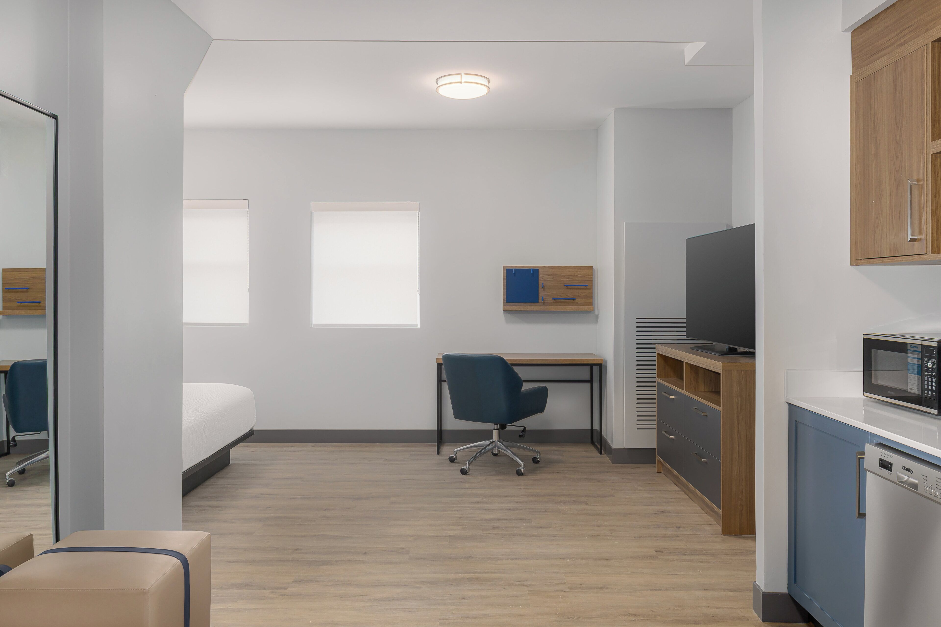 Habitación de hotel pequeña con cama, escritorio, silla, televisor, microondas y un mueble bajo.
