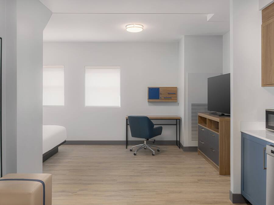 Habitación de hotel pequeña con cama, escritorio, silla, televisor, microondas y un mueble bajo.