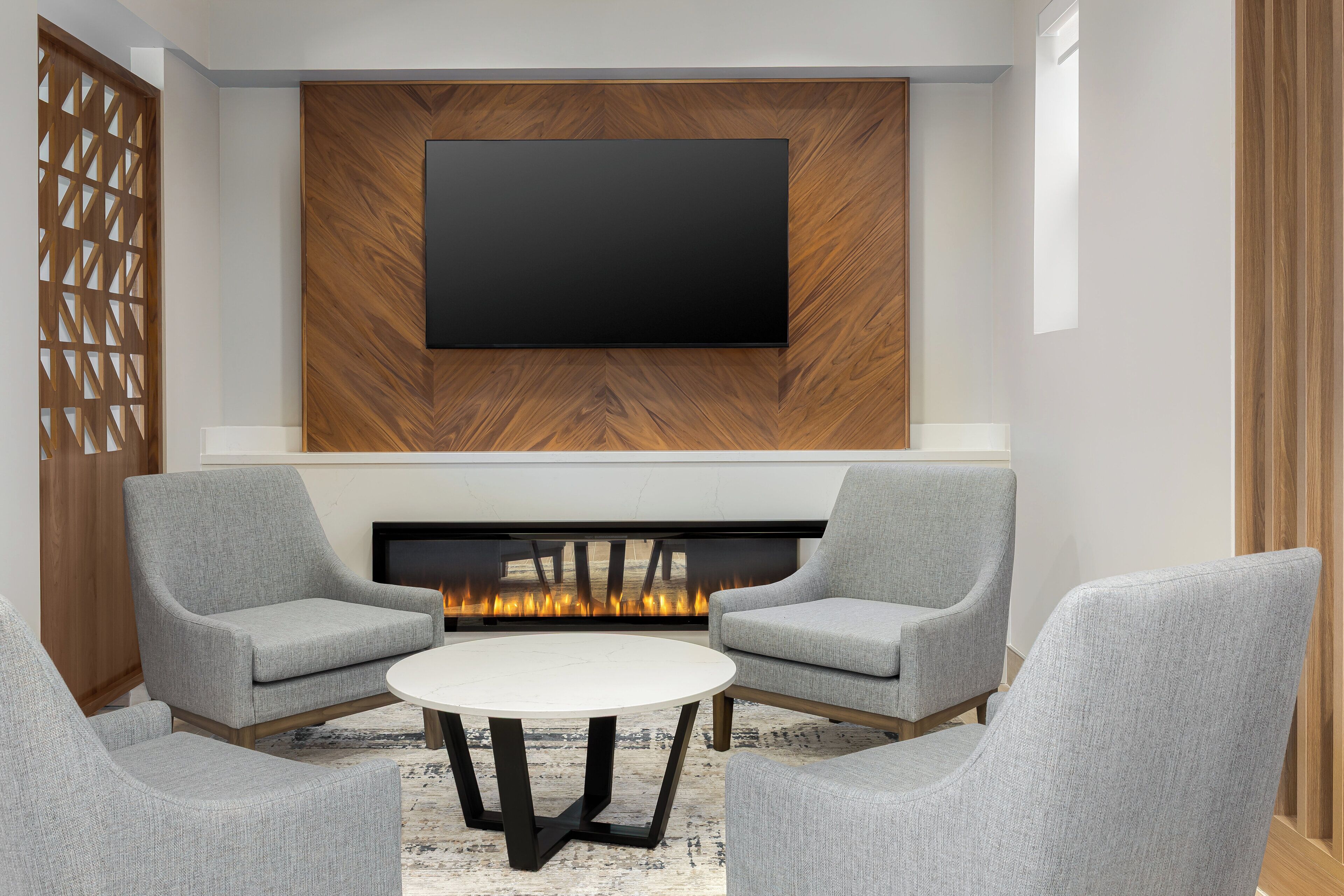 A modern living room with a flat-screen TV mounted on a wooden wall, a fireplace, and grey upholstered chairs around a round coffee table.