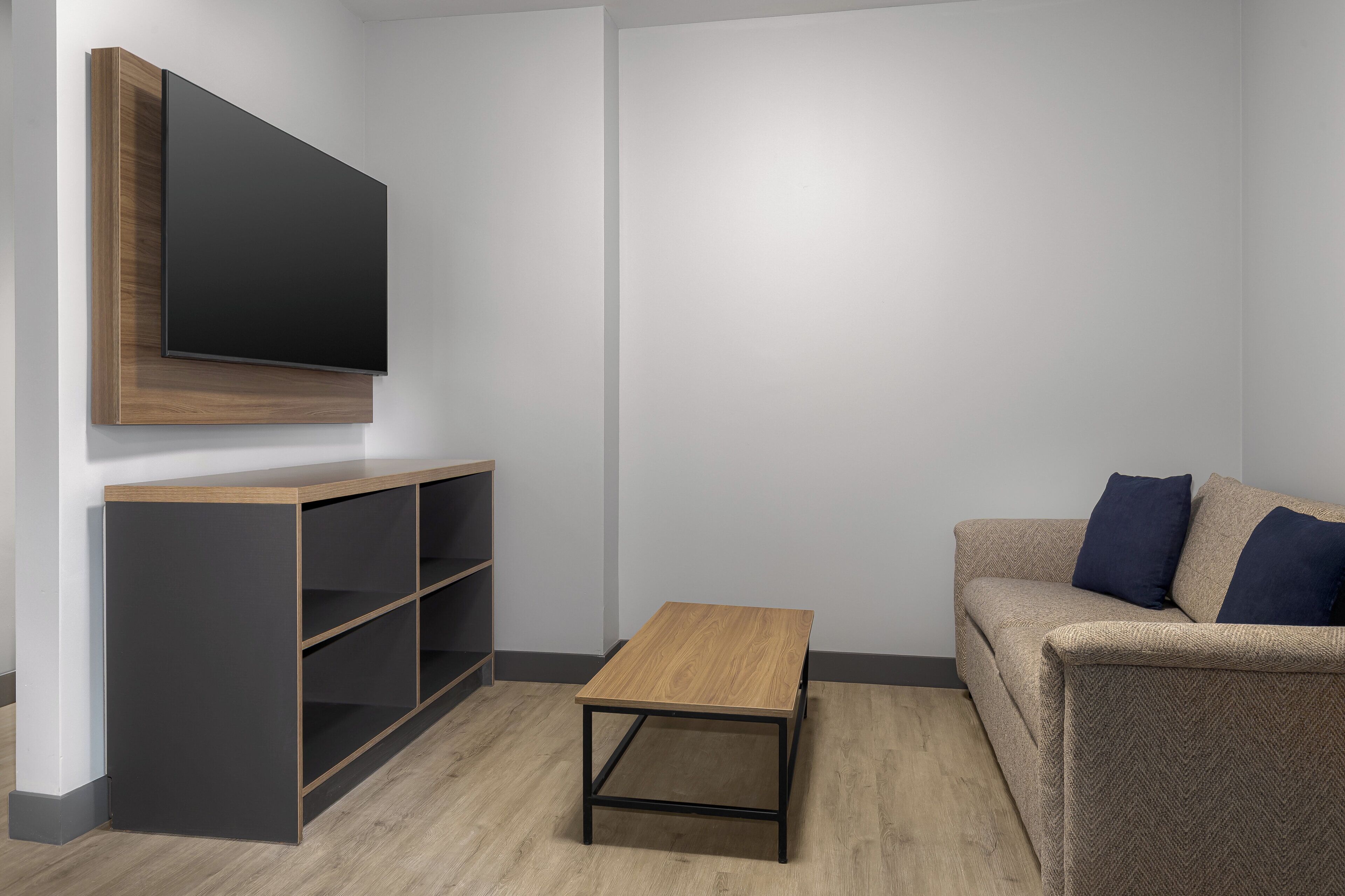Una sala de estar moderna con un televisor de pantalla plana montado en un mueble de pared de madera, una pequeña mesa de centro de madera y un sofá con cojines oscuros.