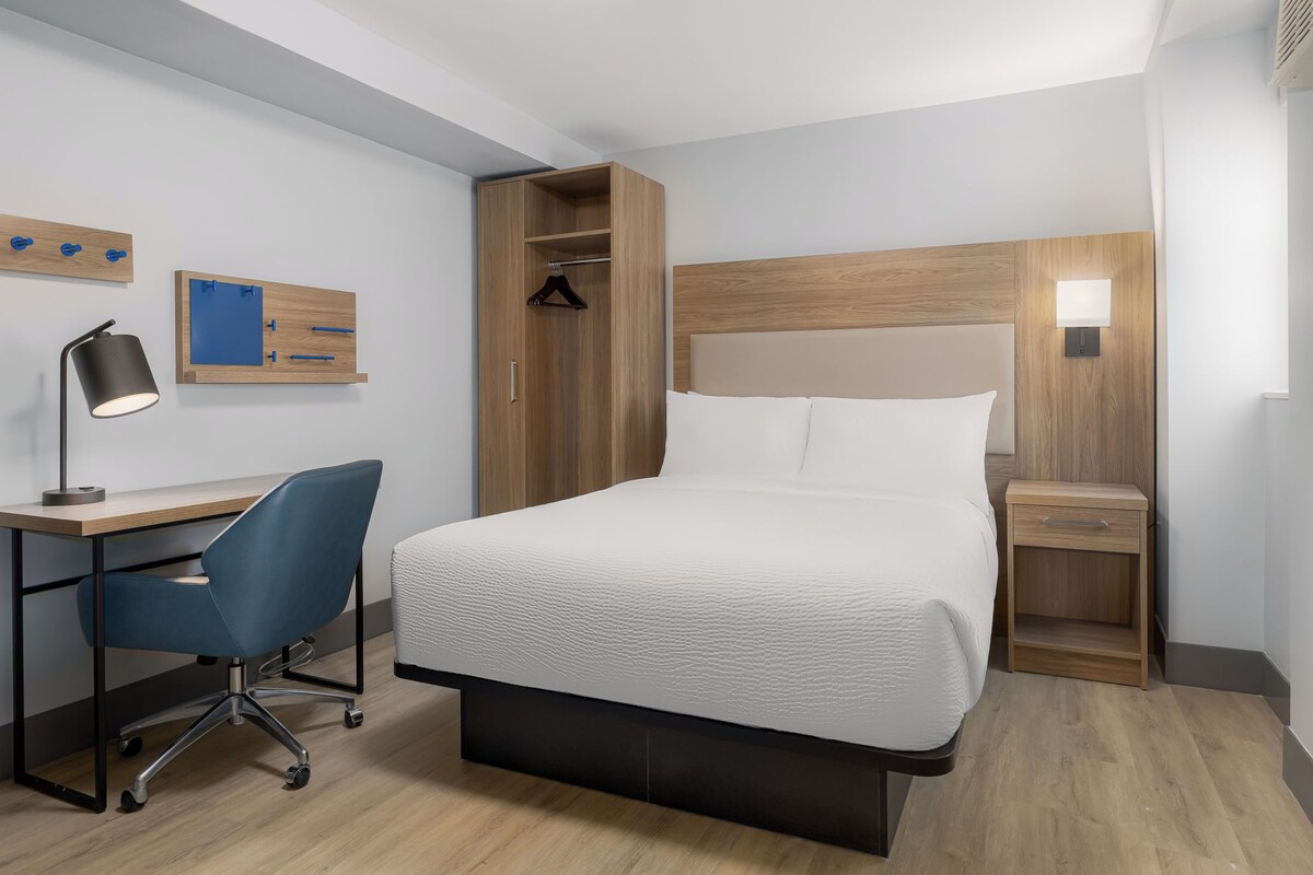 Quarto de hotel com cama, escrivaninha, cadeira, guarda-roupa e mesa de cabeceira.