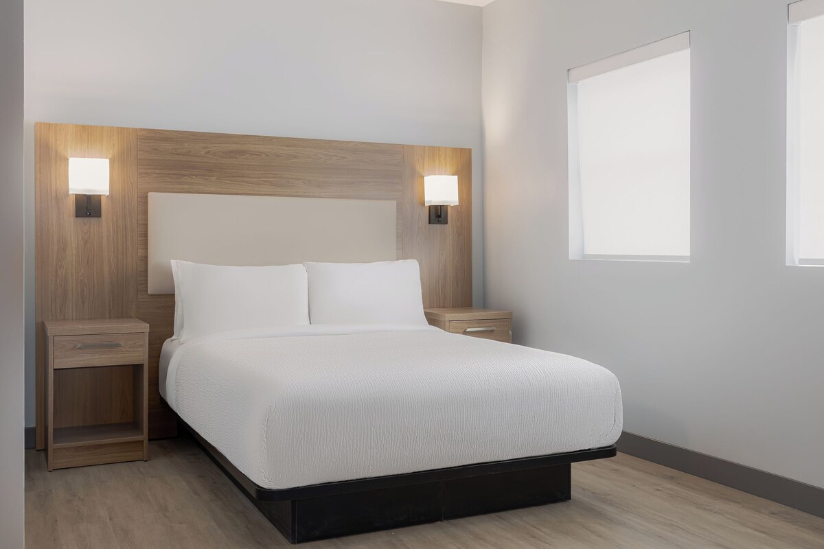 Quarto moderno com cabeceira de madeira, duas mesas de cabeceira, cama com roupa de cama branca e duas luminárias fixadas na parede.