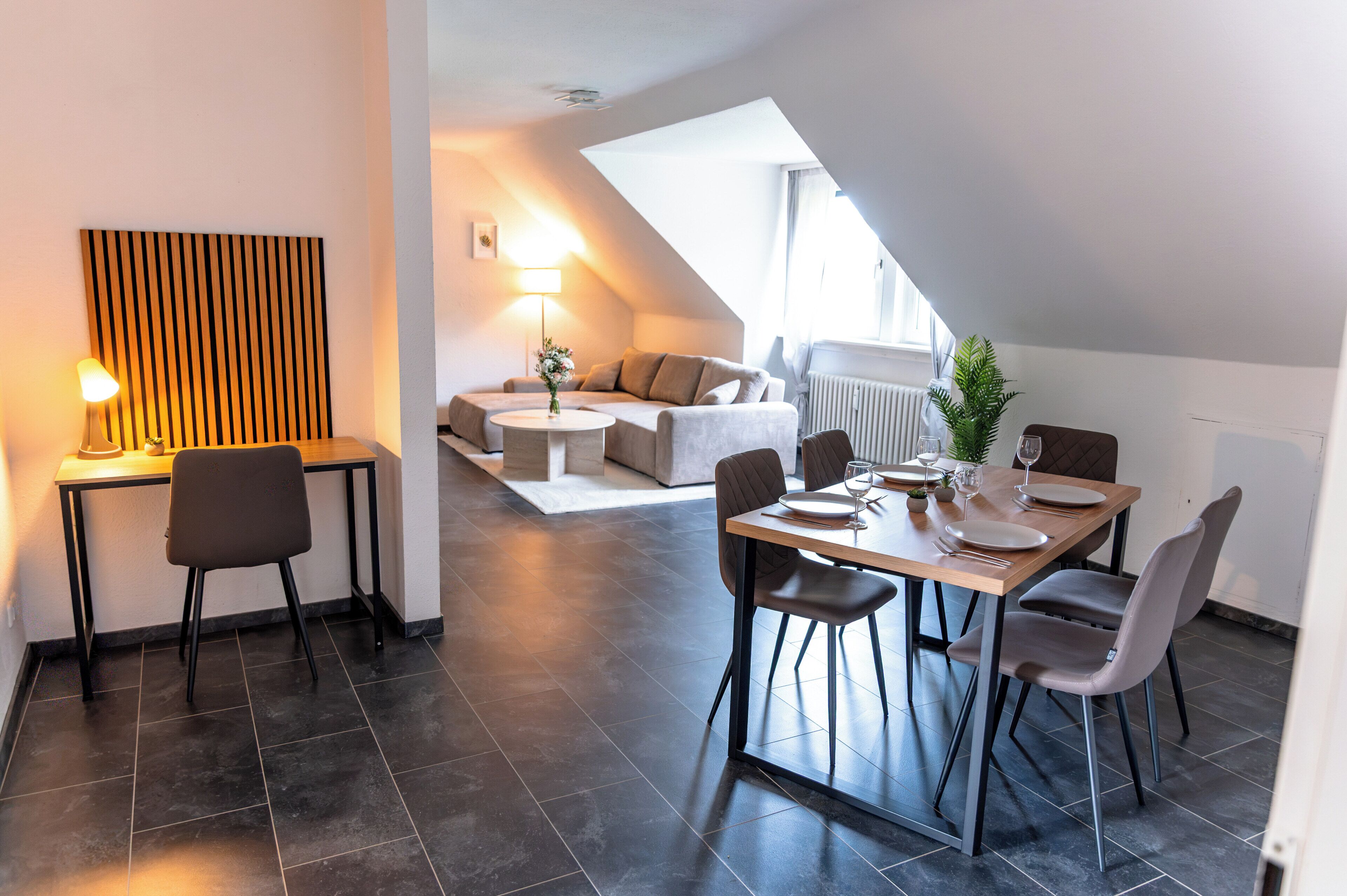 A modern dining area with a table set for two, a sofa in the background, and a lamp on a side table.