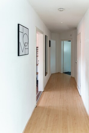 Un pasillo con piso de madera, paredes blancas y una puerta que da a otra habitación.