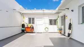 Ett takförsett utomhusområde med ett klinkergolv, en surfbräda lutad mot väggen och en krukväxt nära ingången.