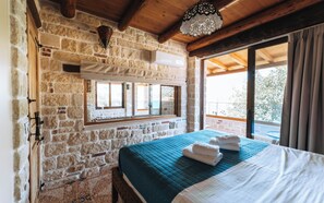 Una habitación con paredes de piedra, una cama con colcha azul, una ventana con vistas y una lámpara colgante.
