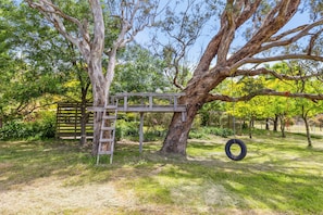 Une cabane dans les arbres, accessible par une Ă©chelle, avec une balançoire et une balançoire faite dâun pneu, le tout situĂ© dans un espace verdoyant bordĂ© dâarbres.