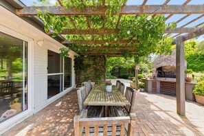 Un patio cubierto con una mesa y sillas de madera, un horno de ladrillo y una pérgola con plantas trepadoras.