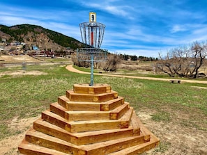 Ein Discgolf-Korb mit einer hölzernen Treppe, die zu ihm hinaufführt, umgeben von einem Rasenfeld und einer hügeligen Landschaft im Hintergrund.
