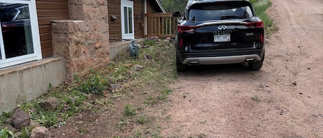 A black SUV parked in front of a house with a stone chimney, a wooden deck, and a dirt path leading to a forested area.
