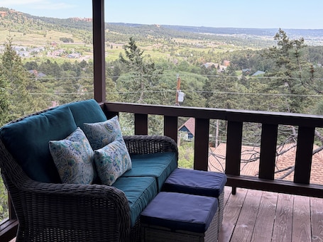 A wicker sofa with cushions on a wooden deck with a view of a rural landscape.. Built in 1908 this historic cabin is full of original charm with a penthouse view. Step inside and you are immediately greeted by a full unobstructed view of Elephant Rock. Sleeping up to 9 people.