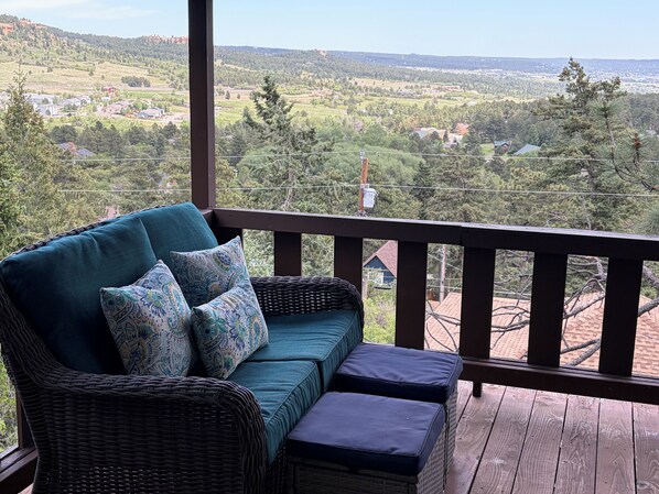 A wicker sofa with cushions on a wooden deck with a view of a rural landscape.