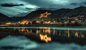 Nachtlandschaft mit einer sternförmigen Lichtinstallation auf einem Hügel, ruhigem Gewässer im Vordergrund und einer kleinen Siedlung mit Häusern und Lichtern im Hintergrund.