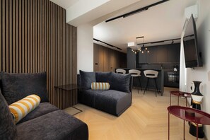 Ein modernes Wohnzimmer mit dunklen Sofas, einer Wandpaneele aus Holz, einer Essecke mit Barhocker und einer Küche mit Fernseher.