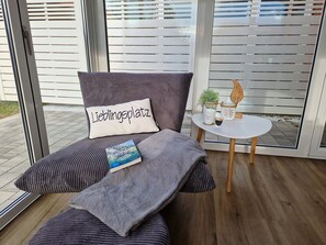 Ein modernes Zimmer mit einer Couch, einem Kissen mit Schriftzug, einem Buch, einem kleinen Tisch mit Pflanze und Tasse sowie Blick durch große Fenster.