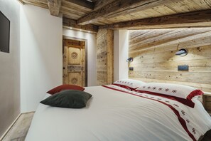 Ein Schlafzimmer mit Holzbalkendecke, einem Bett mit weißer Bettwäsche und roten Akzenten, einer Holztür und einer kleinen Wandleuchte.