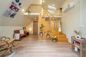 A spacious living area with a wooden staircase, a sofa, a wicker chair, and potted plants. - Lolos by Raho Cozy Loft Stay in Coorg (Virajpet)
