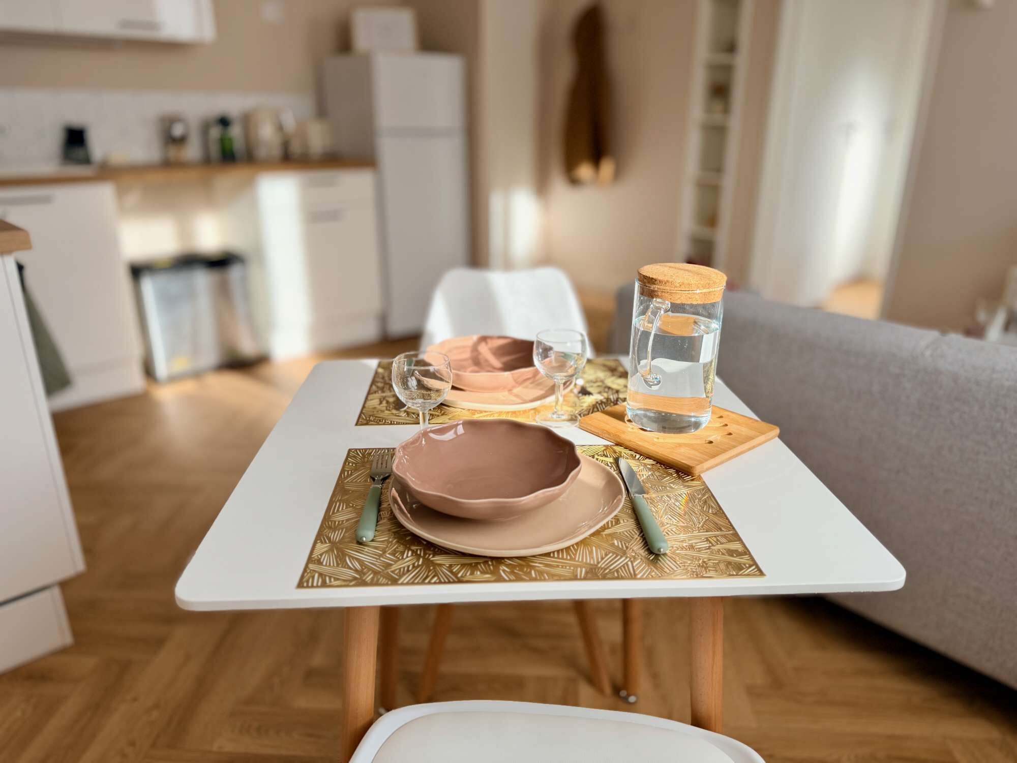 Une table à manger recouverte d’une nappe blanche, avec un bol rose, un verre à vin et une bouteille d’eau posés sur un set de table à motifs.