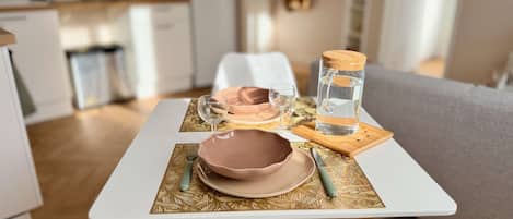 Une table à manger recouverte d’une nappe blanche, avec un bol rose, un verre à vin et une bouteille d’eau posés sur un set de table à motifs.