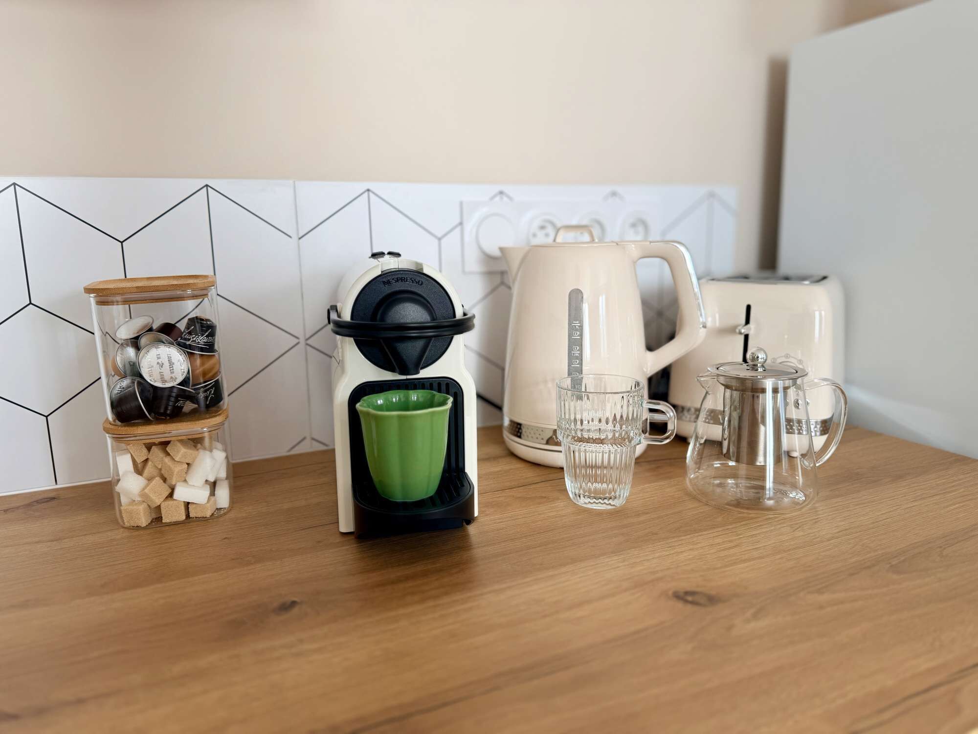 Eine Küchenarbeitsfläche mit Kaffeemaschine, einem Glasgefäß mit Zuckerwürfeln, einer grünen Tasse und einem Teekessel.