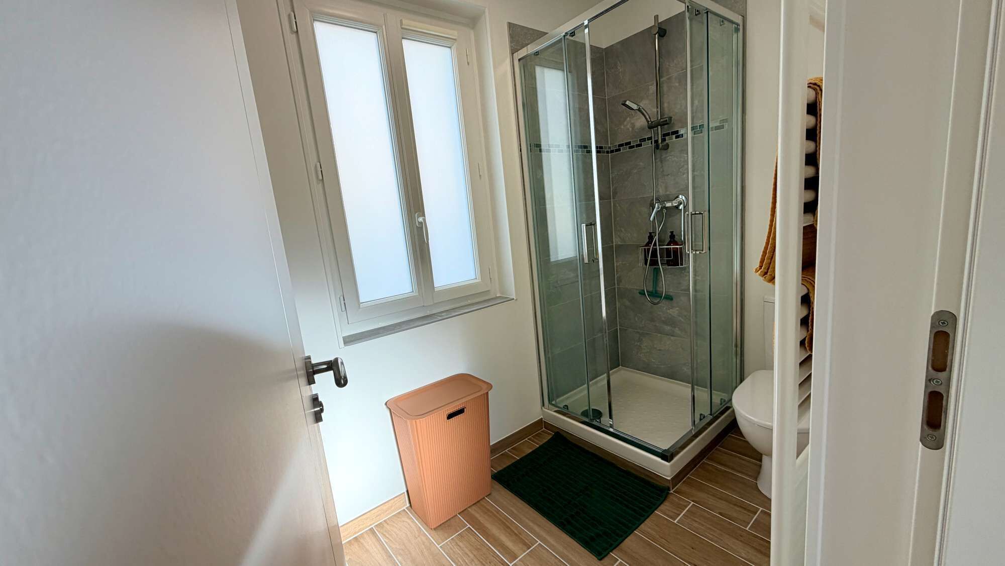 Une salle de bain avec une douche à parois de verre, des toilettes, une poubelle et une fenêtre.