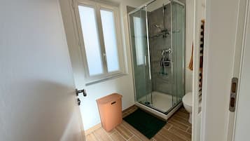 Une salle de bain avec une douche à parois de verre, des toilettes, une poubelle et une fenêtre.