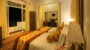 A hotel room with two beds, a desk, a chair, a mirror, and a sliding glass door leading to a balcony. - Queensbury City Hotel - Nuwaraeliya (Nuwaraeliya)
