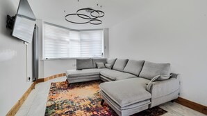 Una sala de estar moderna con un sofá seccional, un tapete colorido, una lámpara de techo y una ventana con persianas.