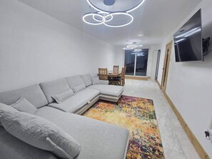 A modern living room with a grey sectional sofa, a colorful rug, a dining area with wooden chairs, and a TV mounted on the wall.