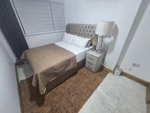 Une chambre à coucher avec un lit, une table de chevet, une lampe, une fenêtre avec des stores et un tapis.