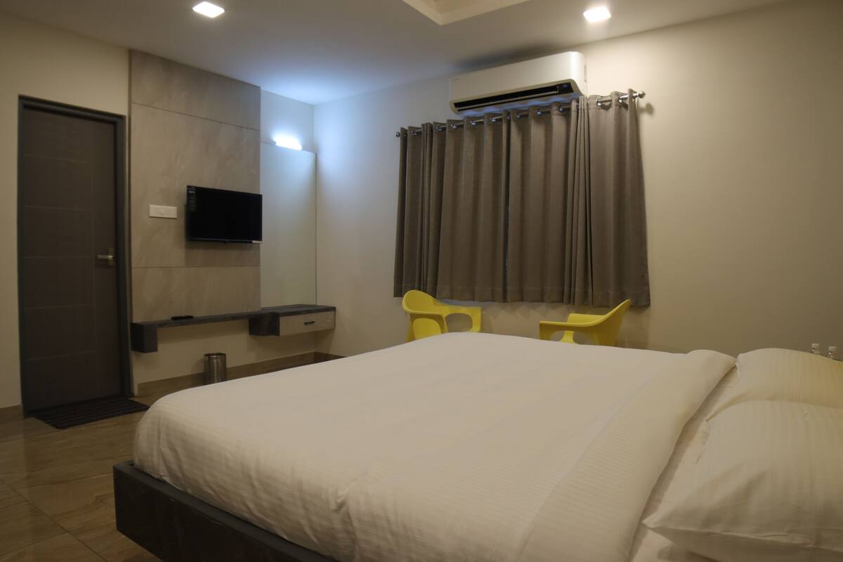 A modern hotel room with a bed, a flat-screen TV mounted on the wall, a desk with a chair, and an air conditioning unit.