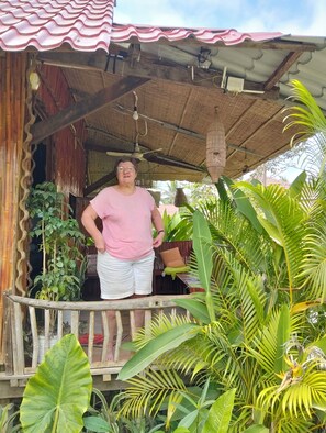 A person standing on a porch with a thatched roof, surrounded by lush tropical plants.