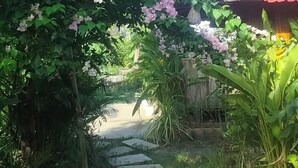 Een tuinpad leidt naar een huis met een rood dak, omgeven door weelderig groen en bloeiende planten.