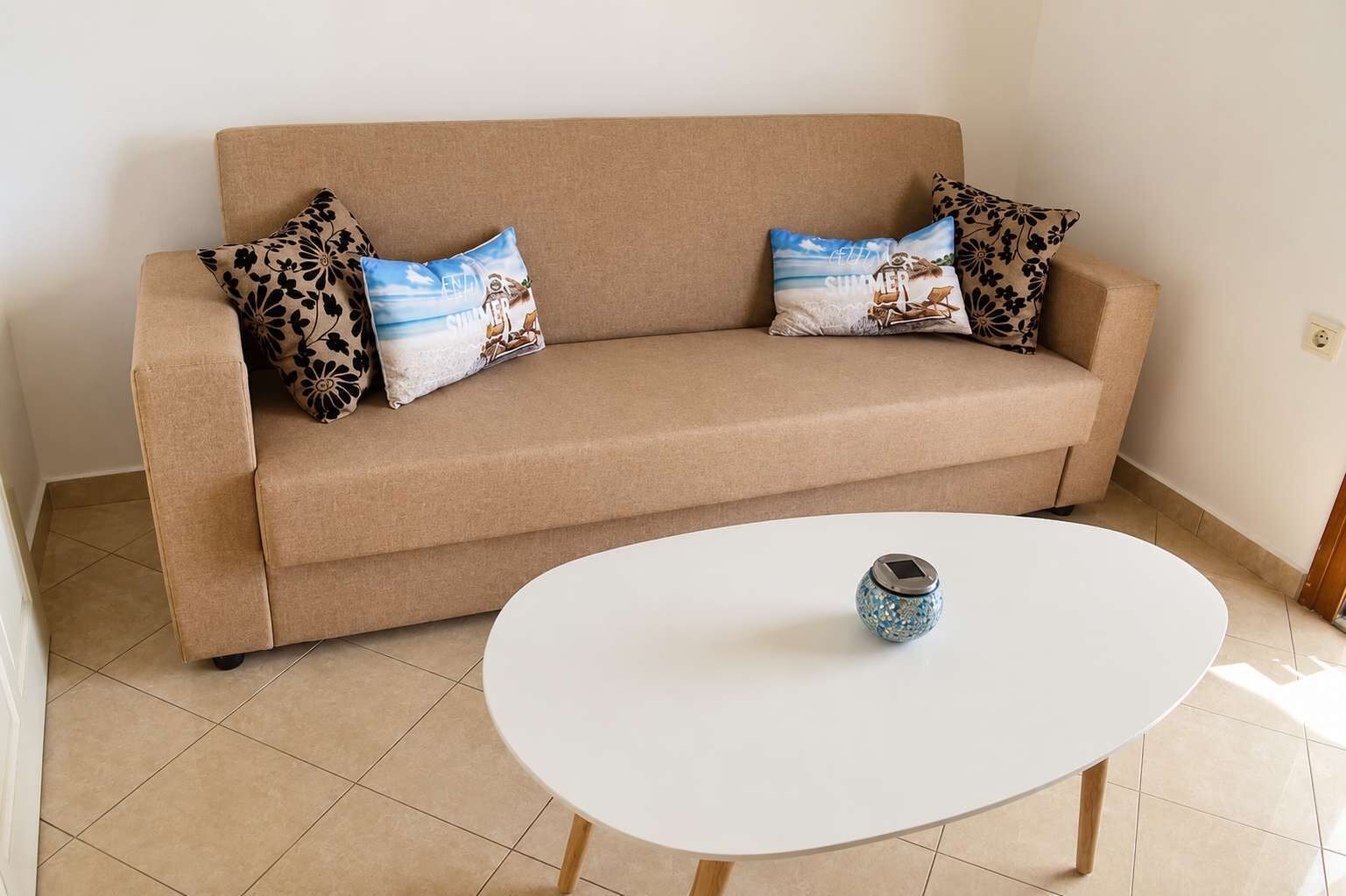 Een beige bank met kussens met bloemen en strandmotieven, een witte tafel met een decoratieve vaas en een betegelde vloer.
