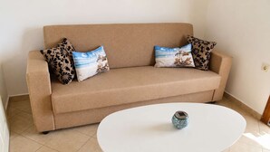 Ein beiges Sofa mit floralen und strandbezogenen Kissen, ein weißer Tisch mit einer Dekorationsvase und ein gefliester Boden.