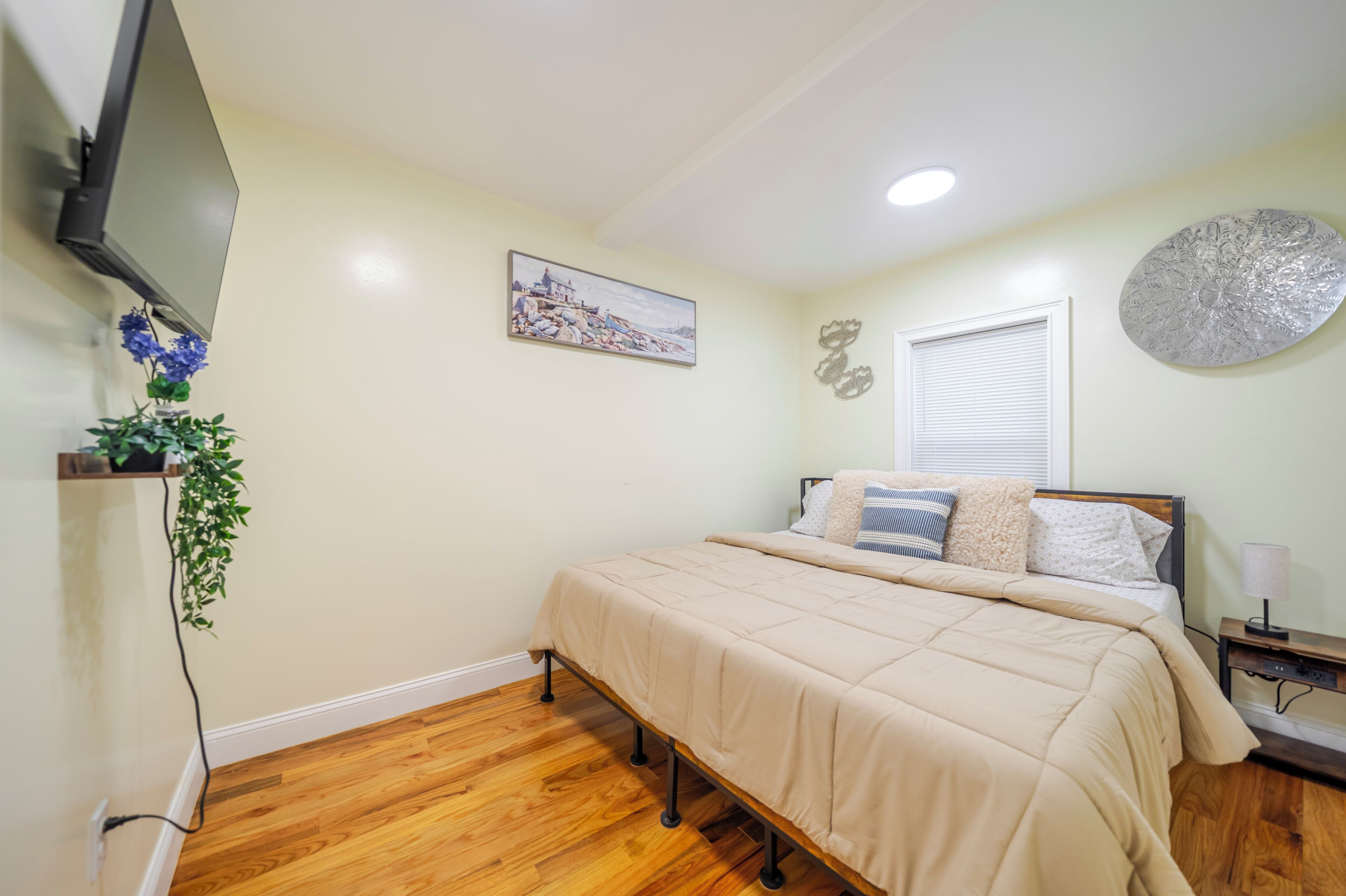 A bedroom with a bed, a TV, a painting, a window, and a decorative wall piece.