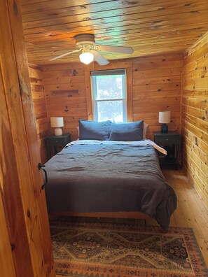 A wooden cabin bedroom with a bed, two bedside tables with lamps, a ceiling fan, and a window.