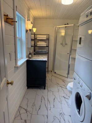 A compact bathroom with a built-in washing machine, a glass shower enclosure, and a vanity with shelves for towels.