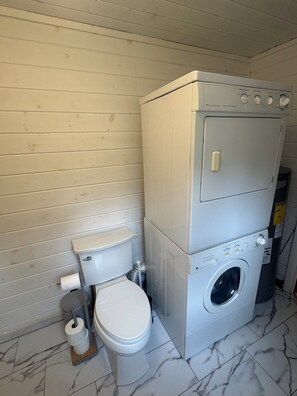 A compact laundry room with a washing machine and dryer stacked on top of each other, a toilet, and a roll of toilet paper.