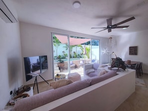 Ein modernes Wohnzimmer mit großem Fernseher, Deckenventilator und einer Schiebeglastür, die auf eine Terrasse im Freien führt.