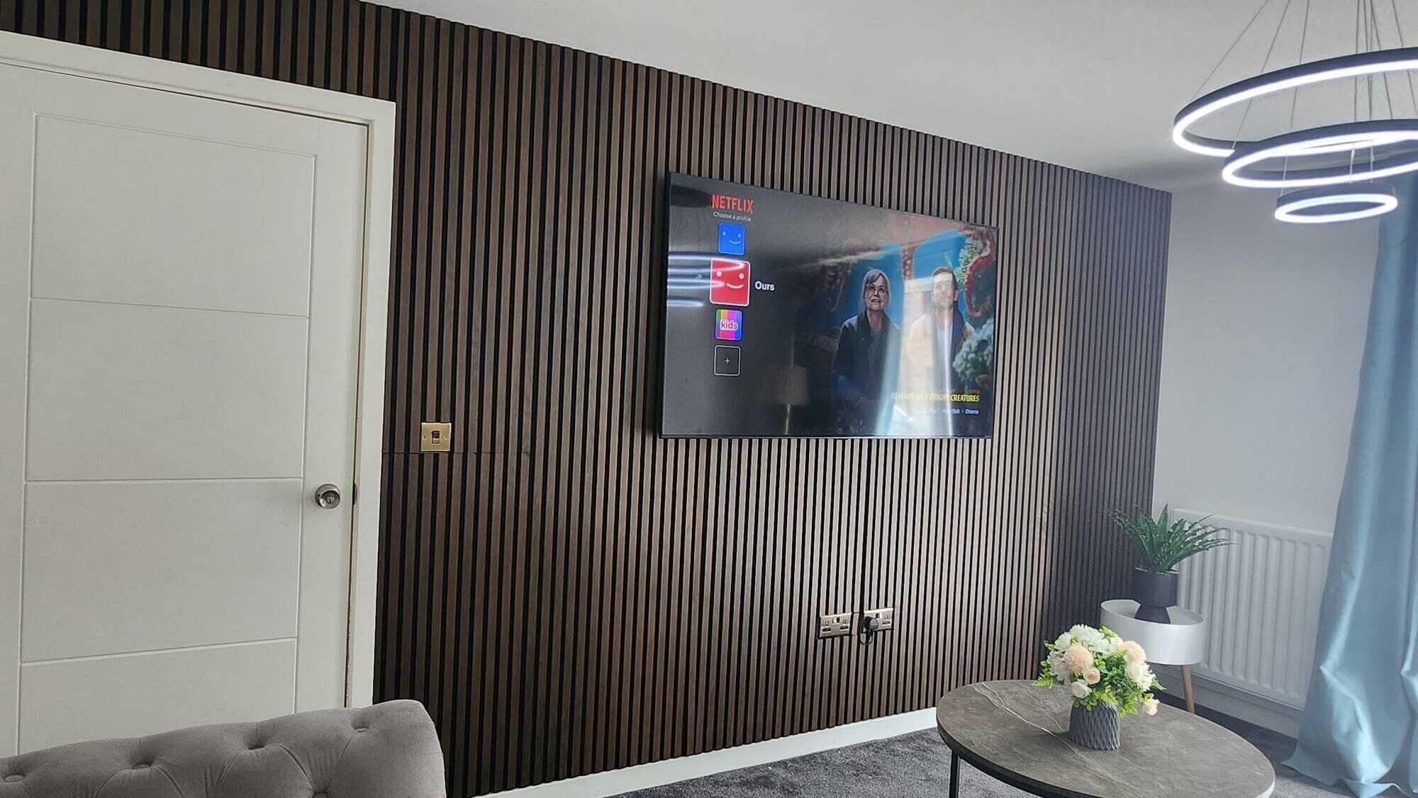 Un salon moderne avec un téléviseur à écran plat fixé à un mur en bois, un canapé gris, une petite table ronde ornée de fleurs et un luminaire suspendu au plafond.