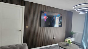 Ein modernes Wohnzimmer mit einem an einer Holzwand montierten Flachbildfernseher, einem grauen Sofa, einem kleinen runden Tisch mit Blumen und einer Deckenleuchte.